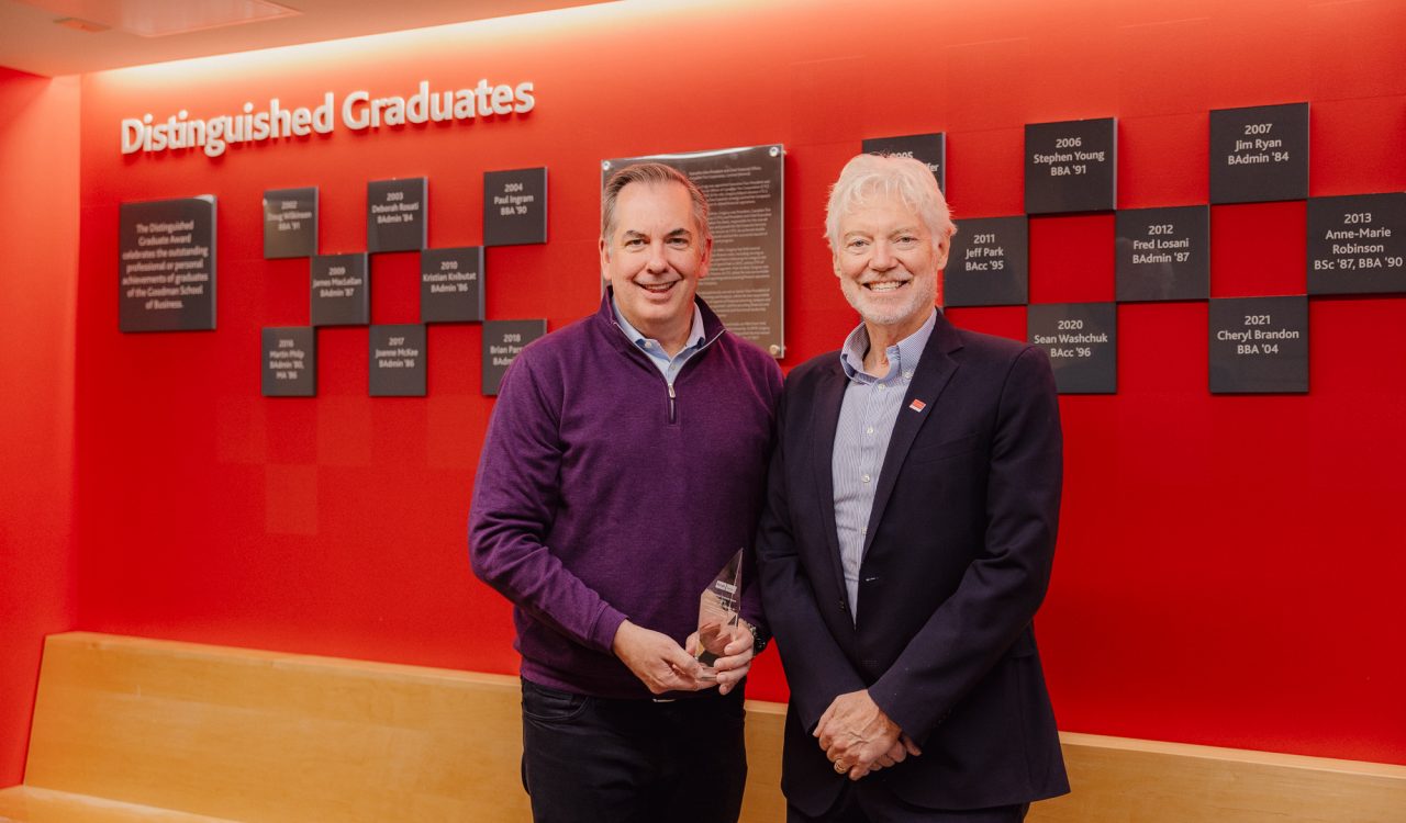 Two men stand in front of a red wall that reads Distinguished Graduates with small grey plaques. The man on the left holds a glass trophy.