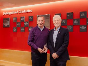 Two men stand in front of a red wall that reads Distinguished Graduates with small grey plaques. The man on the left holds a glass trophy.