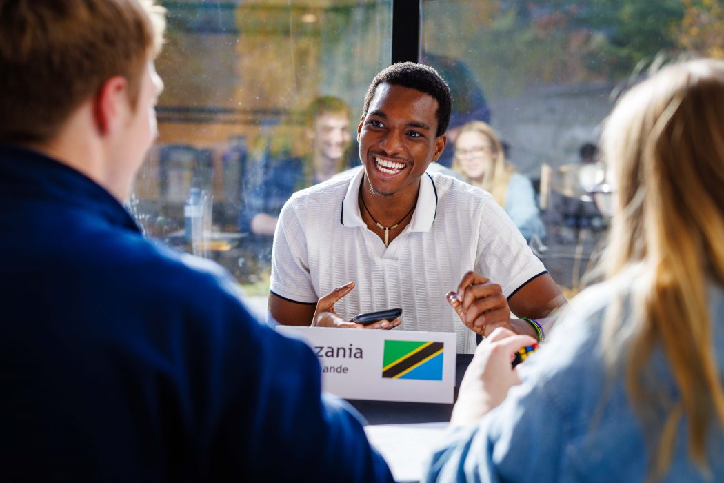 A smiling young man sits at a table with two other people, engaging in a friendly conversation.
