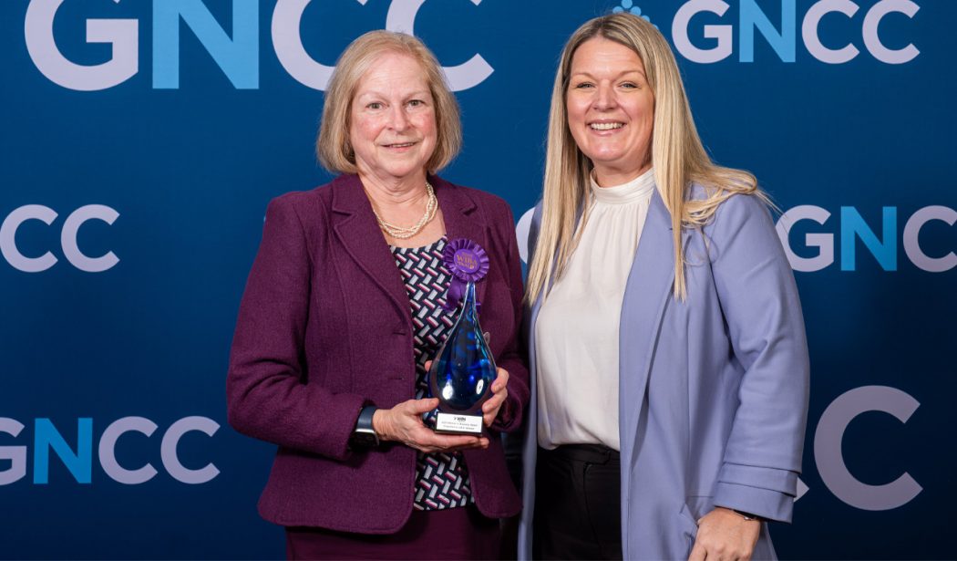 Two women pose with an award in front of a blue backdrop.