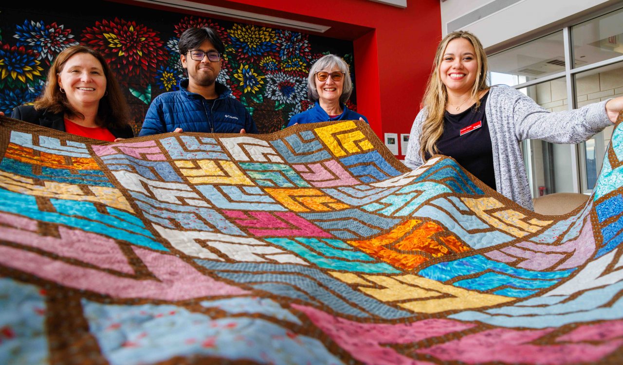Four smiling people hold up a large, colourful quilt featuring geometric patchwork patterns in blue, pink, orange and yellow tones inside a bright room with red walls and a floral mural in the background.