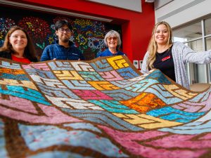 Four smiling people hold up a large, colourful quilt featuring geometric patchwork patterns in blue, pink, orange and yellow tones inside a bright room with red walls and a floral mural in the background.