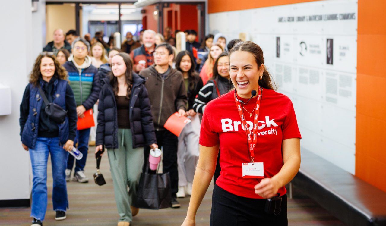 A smiling Brock University student tour guide wearing a red Brock University shirt leads a group of prospective students and families through a bright campus hallway during an open house event.