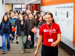 A smiling Brock University student tour guide wearing a red Brock University shirt leads a group of prospective students and families through a bright campus hallway during an open house event.