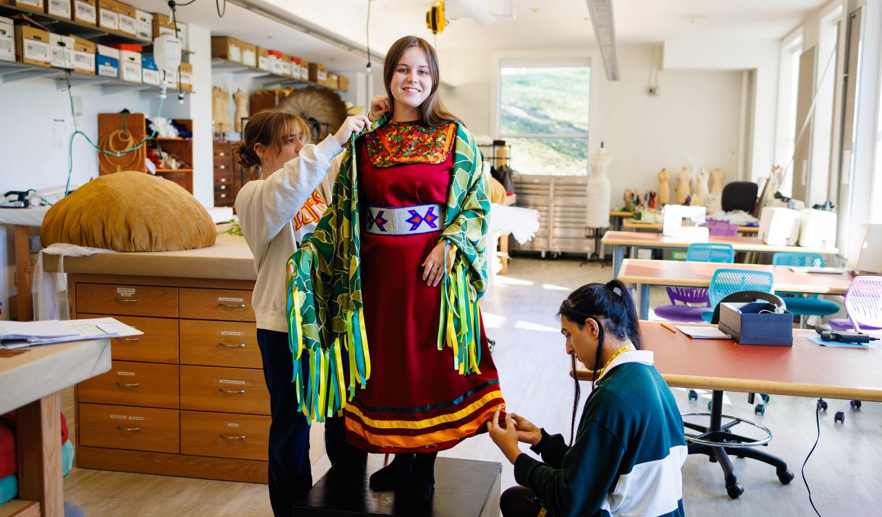 A woman stands on a wood block in a brightly lit wardrobe shop wearing a colourful dress while two people make adjustments to the dress with pins.