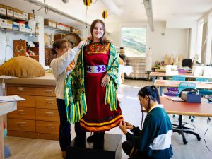 A woman stands on a wood block in a brightly lit wardrobe shop wearing a colourful dress while two people make adjustments to the dress with pins.