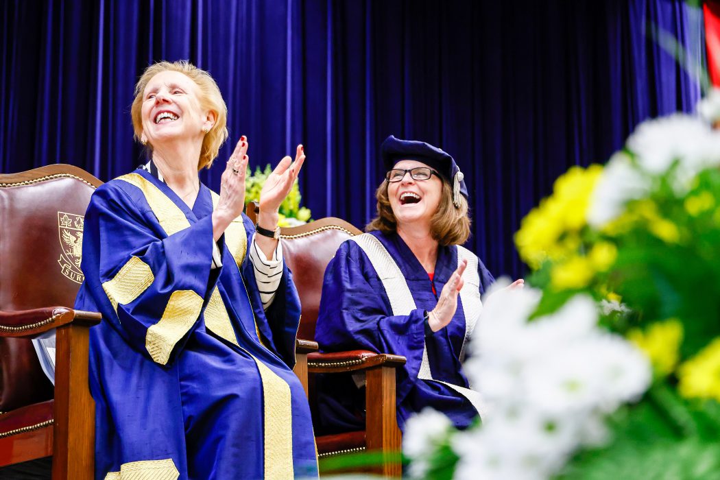 Two women in Convocation gowns clap with excitement as the look on from a stage.