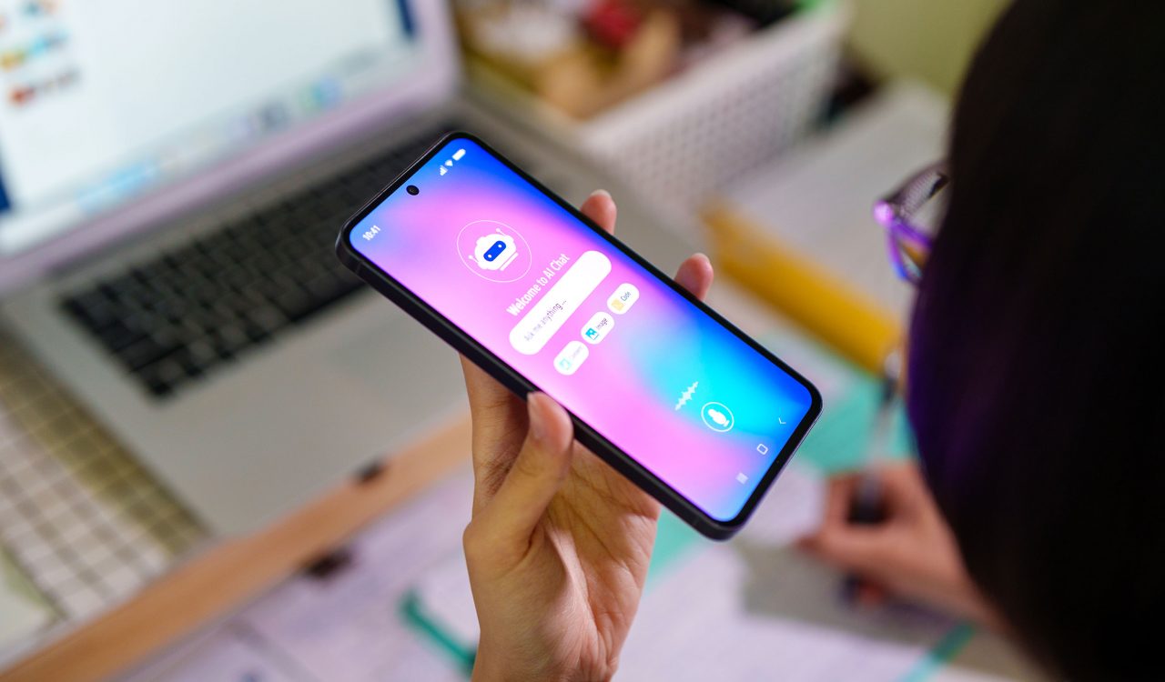Person holds a cell phone using AI while working on a computer.