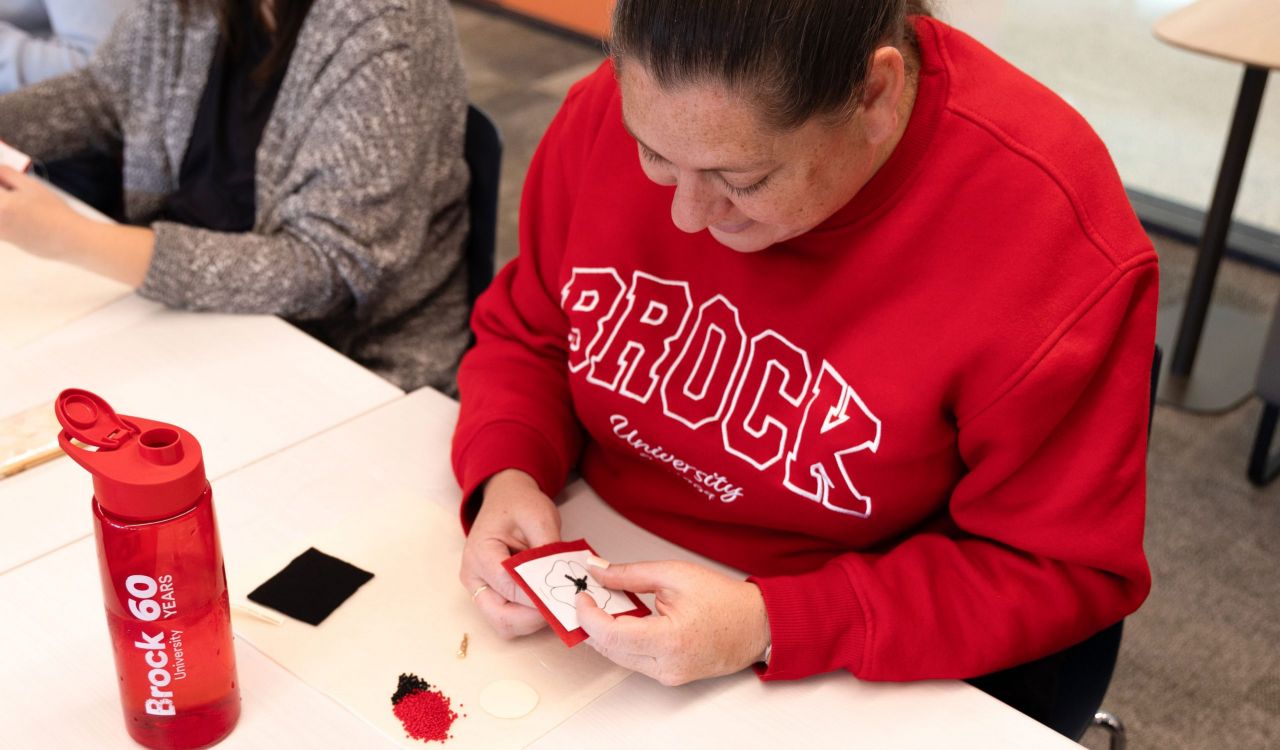 Woman wearing red sweatshirt makes a beaded poppy at a table.