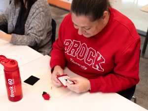 Woman wearing red sweatshirt makes a beaded poppy at a table.