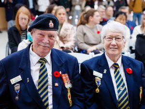 A man and a woman in military uniform sit in the crowd at a Remembrance Day ceremony.