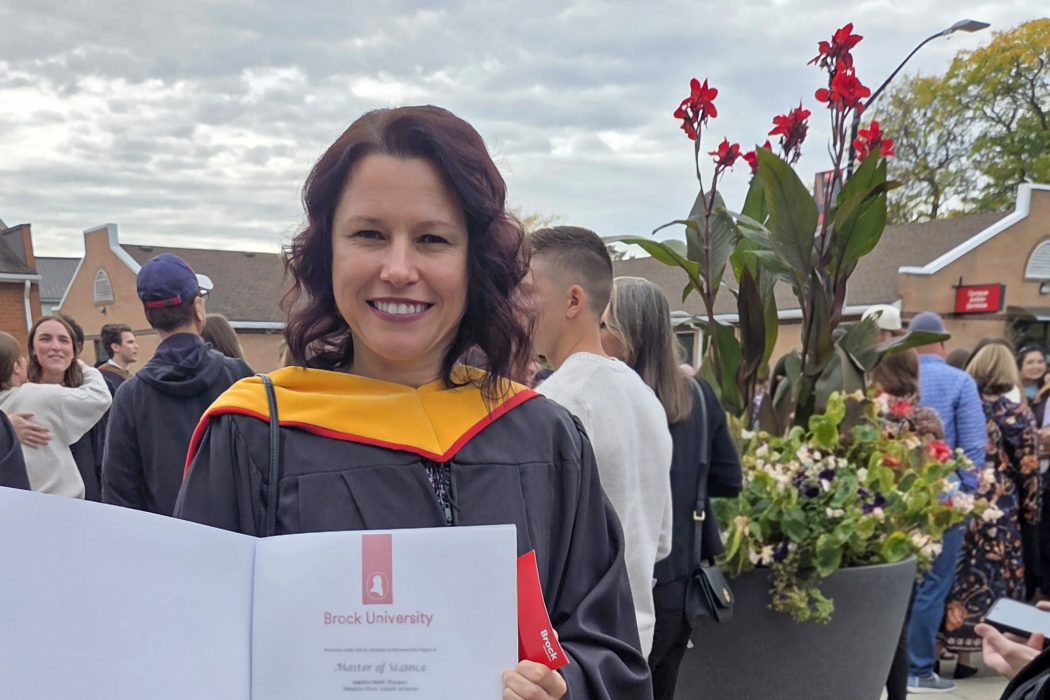A woman in a graduation gown poses with a university diploma.
