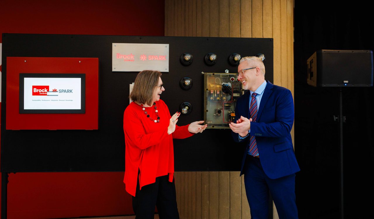 Two people smile and gesture toward each other while unveiling or demonstrating a Brock SPARK display panel.