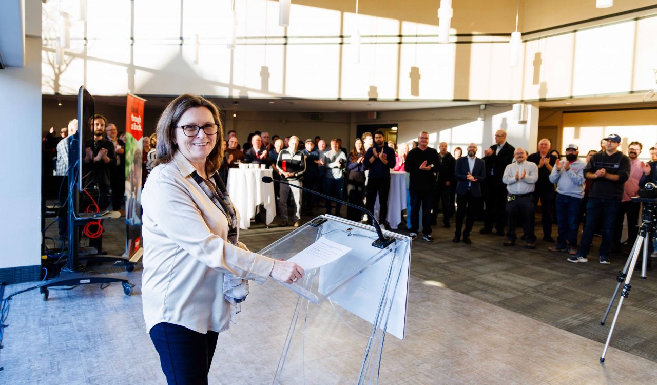 Lesley Rigg smiling at a podium while presenting to a group of Brock faculty and staff gathered in a large brightly lit room an awards celebration.