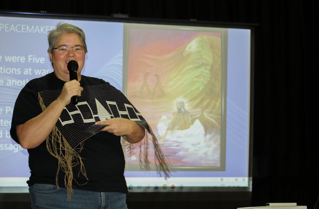 A woman stands holding beaded belt while speaking into a microphone.