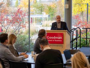 A man in a suit stands behind a podium that reads Goodman School of business. People sit at round tables in the foreground of the image.