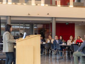 A woman in a blazer speaks at a podium to an audience seated at round tables at an atrium. People are listening attentively, with modern interior in the background.