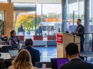 A man wearing blazer stands behind a podium labeled “Goodman School of Business, Brock University” while speaking to attendees seated around tables. Autumn trees, bus, and passing students are visible outside the glass wall.