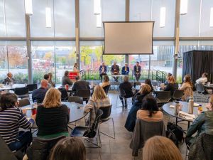A woman in a blazer moderates a panel discussion from a podium labeled “Goodman School of Business” while four panelists sit on stage. The audience watches attentively in a modern, well-lit hall with tall glass windows with fall views.