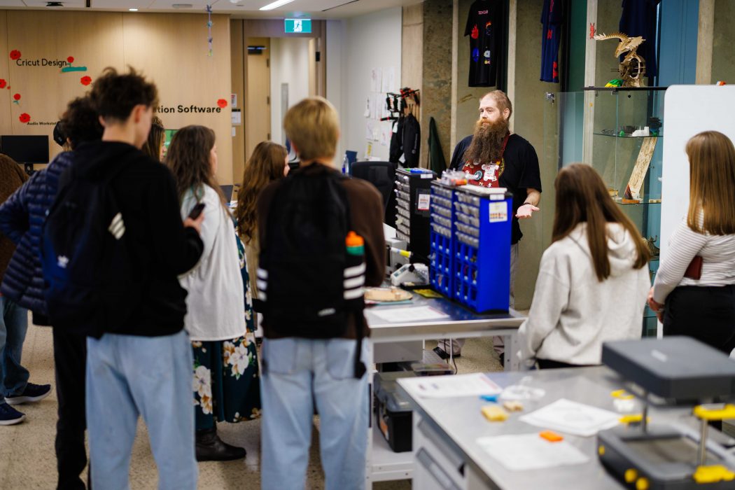 A group of students stand around a workshop table listening to an instructor giving a demonstration. The setting is a makerspace technology lab, with labeled drawers, tools and 3D-printed models on display.