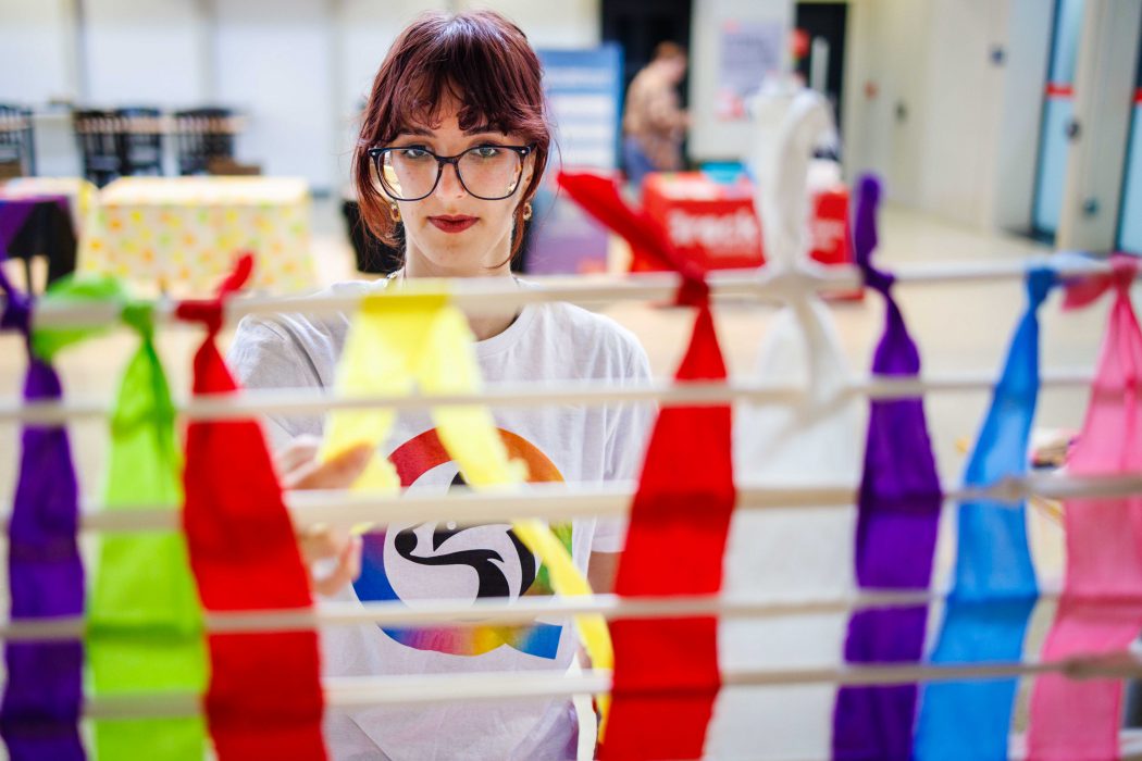 A person wearing a white shirt with a colourful circular logo arranges bright, multicolored ribbons on a rack indoors.