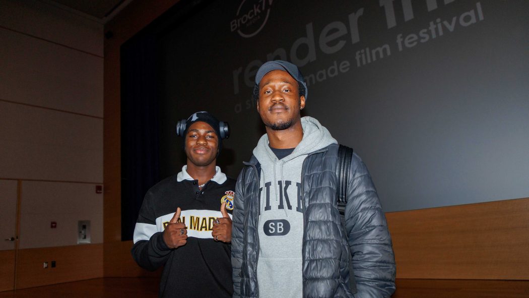 Two university students smile for a photo in front of a large cinema screen.