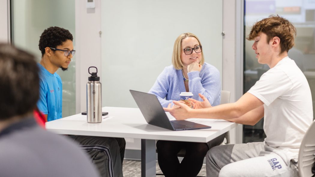 A woman speaks to two university students while sitting at a table.