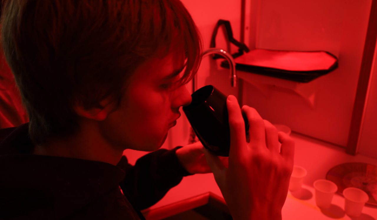 A young man sits at a desk with barriers in front and to the left and right of him under a red light. He puts a glass of liquid to his nose to smell the contents.