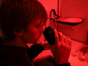 A young man sits at a desk with barriers in front and to the left and right of him under a red light. He puts a glass of liquid to his nose to smell the contents.