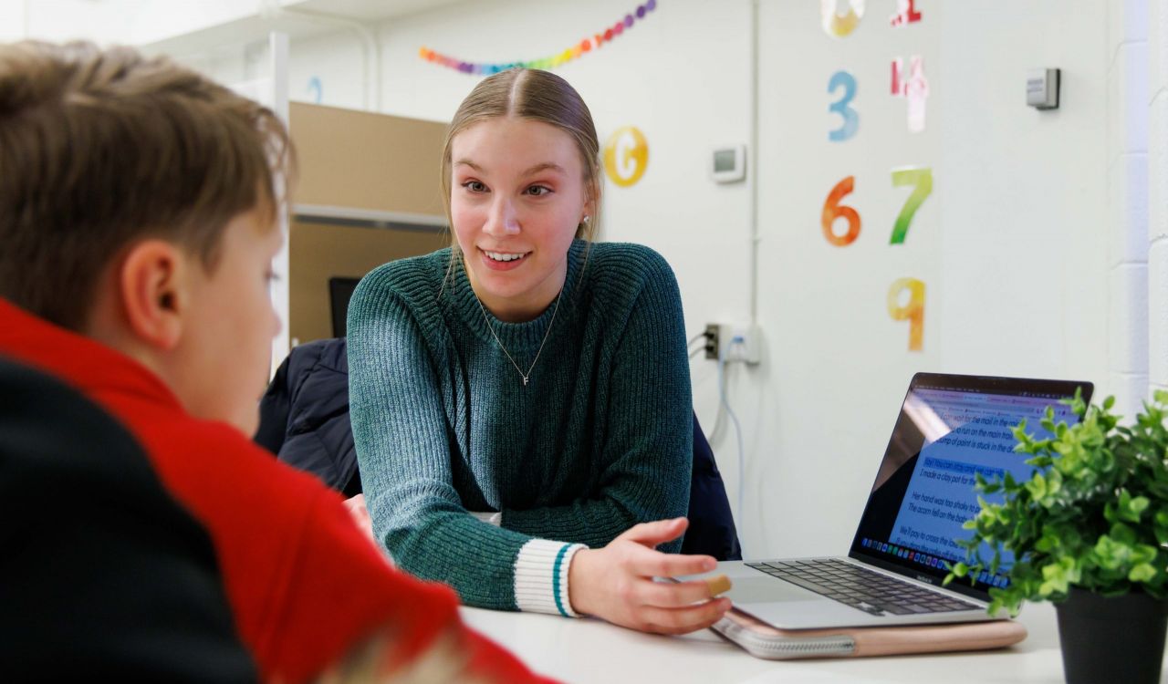 A woman sits across a desk from a child while teaching.