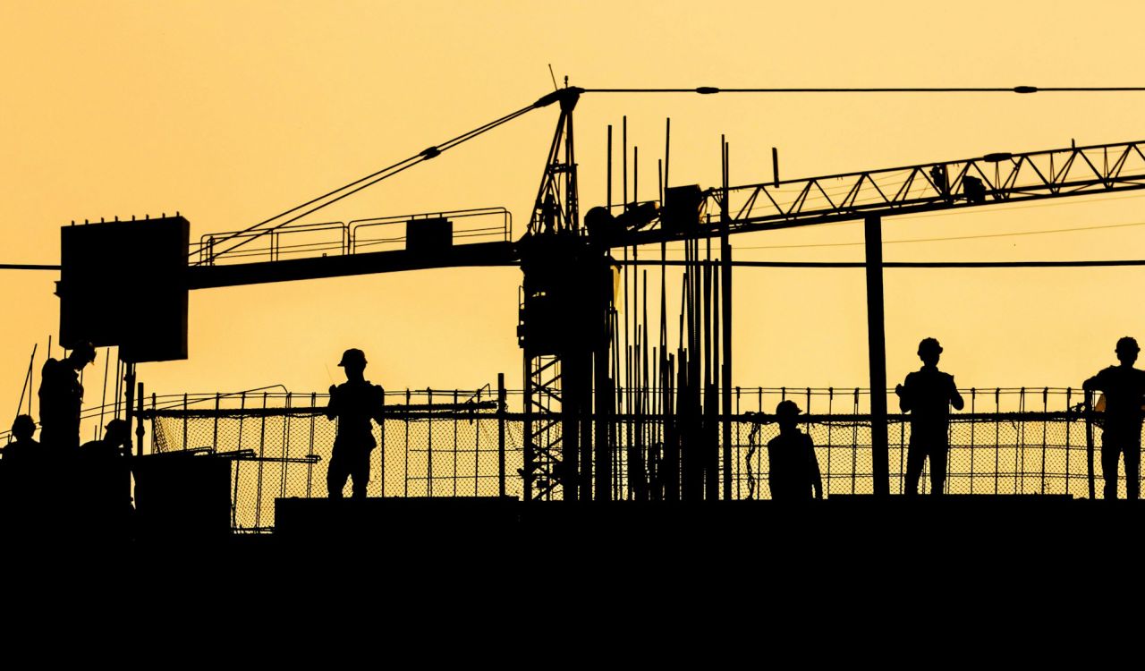 A silhouette of people working on a construction site with a crane and fencing.