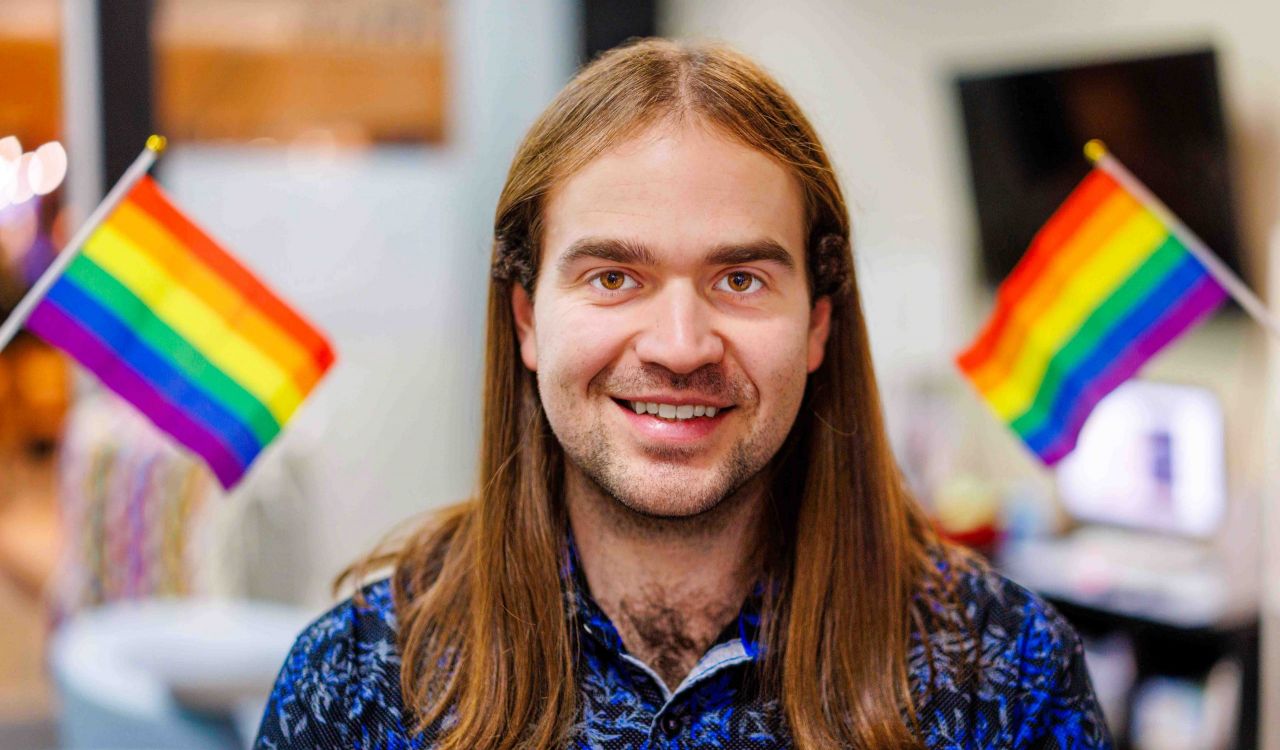 Man wears a blue shirt and is pictured standing beside a rainbow flag.