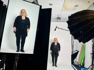 A screen displays the image of Francine McCarthy as she stands in the distance against a white backdrop, with a microphone and a hooded light stand pointing at her.