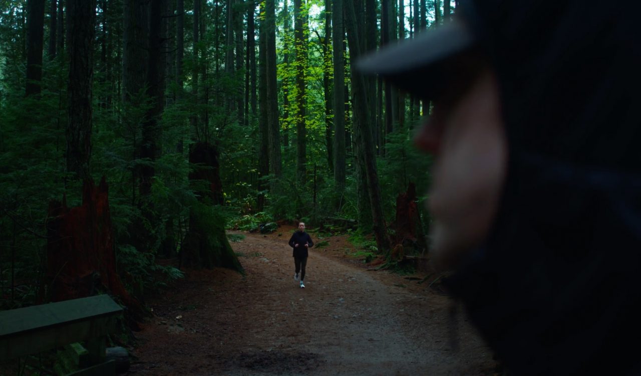 A woman jogging on a path through the woods comes towards a man whose face is seen in the foreground.