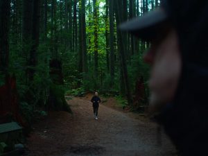 A woman jogging on a path through the woods comes towards a man whose face is seen in the foreground.