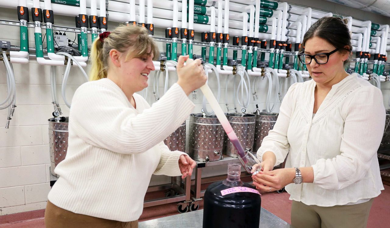 Two women stand in a winery in front of shiny metal fermenting containers and above a jug of test wine while using a long liquid baster to pour wine into a glass.