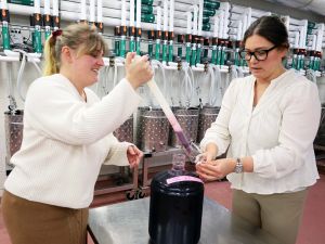 Two women stand in a winery in front of shiny metal fermenting containers and above a jug of test wine while using a long liquid baster to pour wine into a glass.