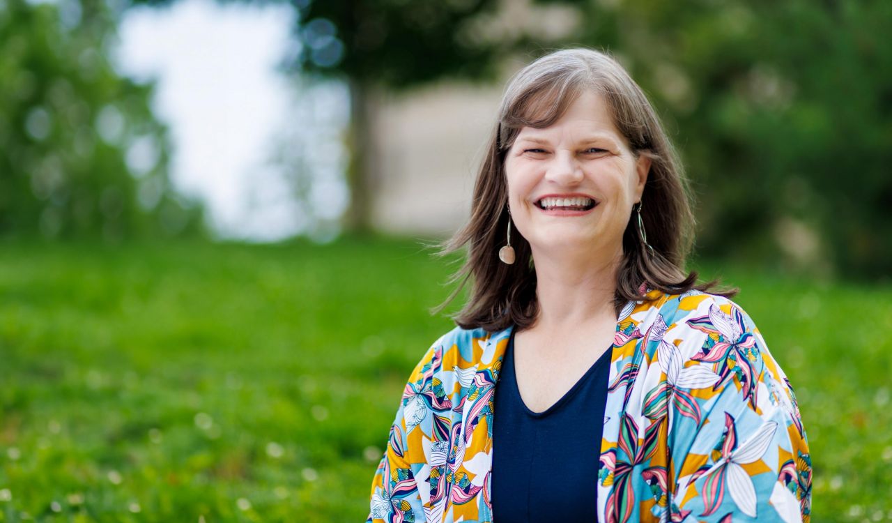 A smiling woman stands outside in a green space.