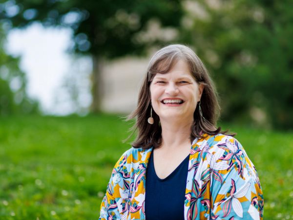 A smiling woman stands outside in a green space.