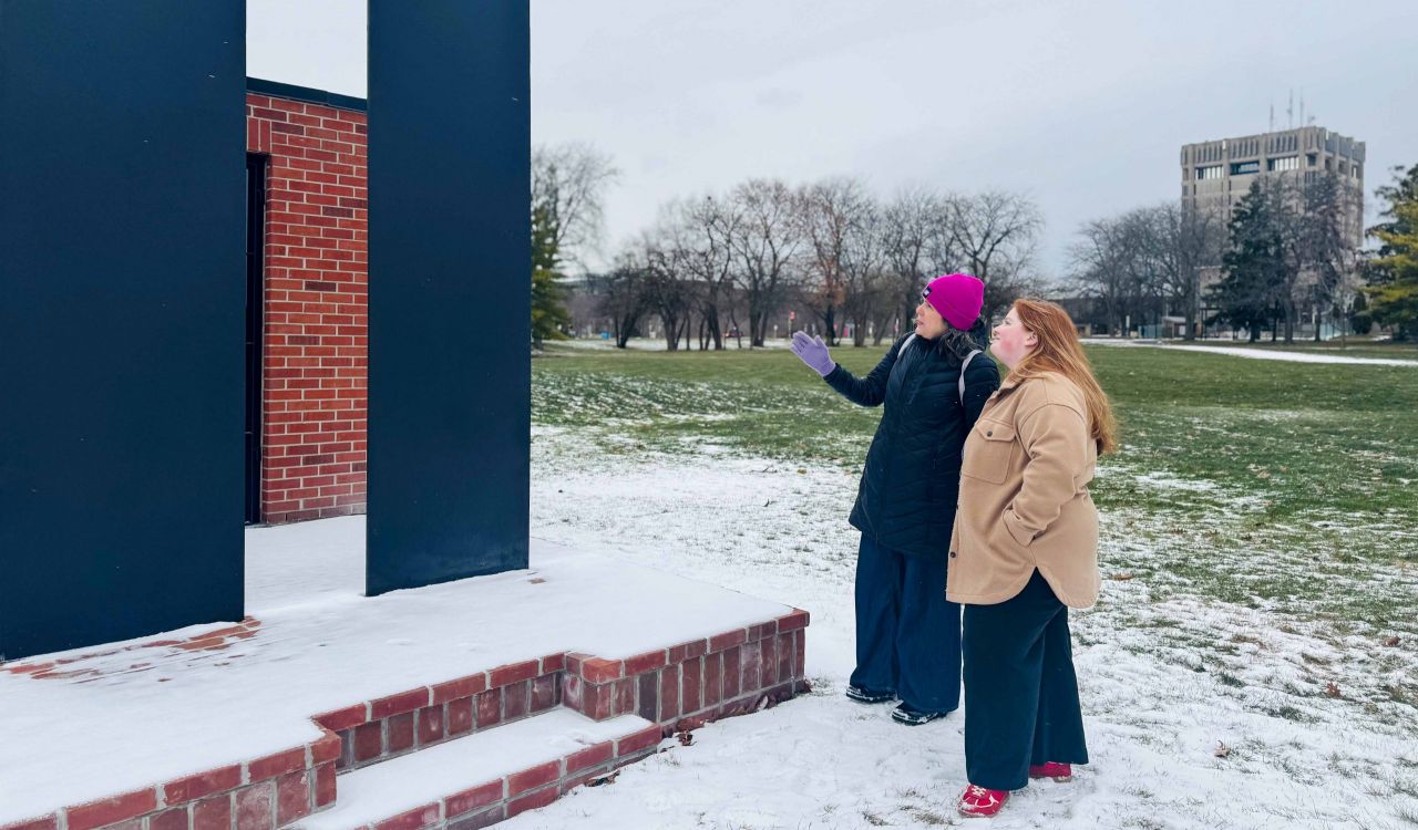 Two women stand outside in winter weather surveying a large sculpture made of brick and steel.