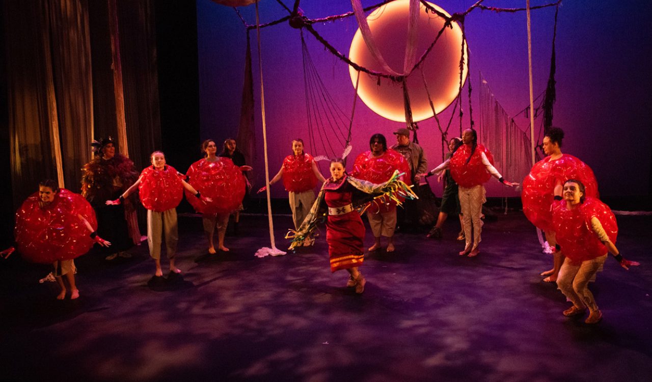 A woman in Indigenous regalia dances on a theatre stage while surrounded by cast members with inflatable costumes around their torsos.