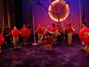 A woman in Indigenous regalia dances on a theatre stage while surrounded by cast members with inflatable costumes around their torsos.