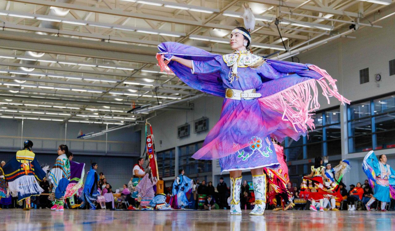 Woman in purple regalia does a traditional dance in a crowded gymnasium.