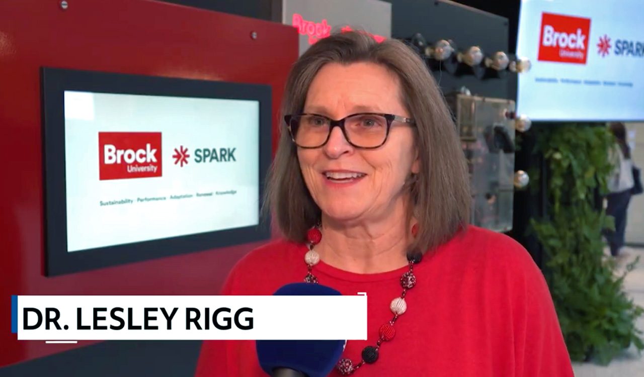 Woman does a television interview in front of a screen that says Brock University and spark.