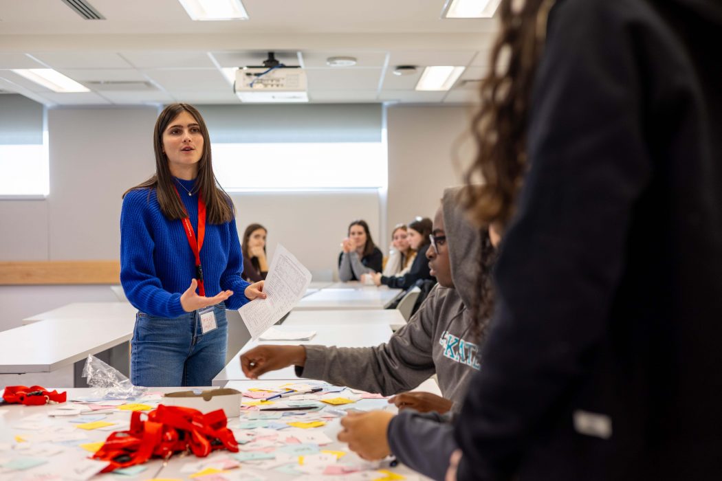 Brock University second-year Speech Language and Hearing Sciences student Mya Booker speaks to a group of students that are seated around a table that is covered in colourful paper squares.