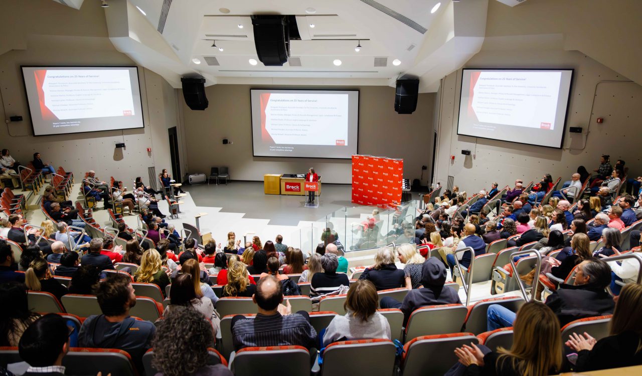 Wide view of a packed lecture hall during a Brock University staff awards event, with a speaker at a podium and presentation slides projected on three large screens.