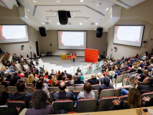 Wide view of a packed lecture hall during a Brock University staff awards event, with a speaker at a podium and presentation slides projected on three large screens.