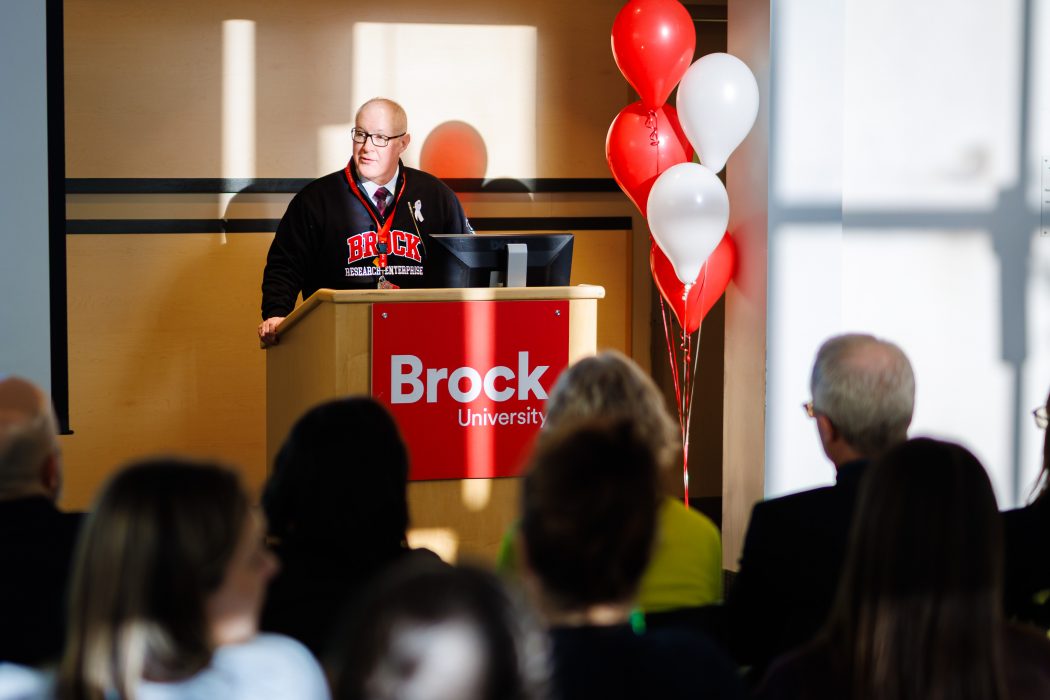 A speaker in a black Brock University sweatshirt stands at a podium with a ‘Brock University’ sign, addressing an audience; red and white balloons are tied beside the stage and sunlight casts strong shadows on the wall.