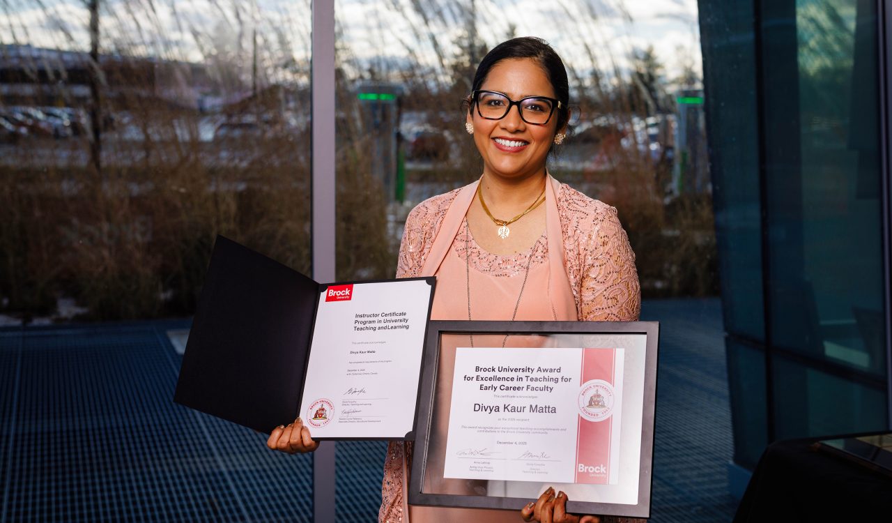 Brock University award recipient Divya Kaur Matta holds an open certificate folder and a framed ‘Excellence in Teaching for Early Career Faculty’ award in front of large windows.