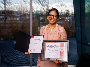 Brock University award recipient Divya Kaur Matta holds an open certificate folder and a framed ‘Excellence in Teaching for Early Career Faculty’ award in front of large windows.